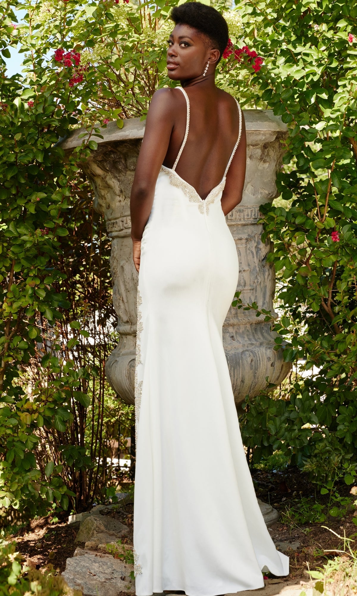 A woman stands outdoors in front of a large stone planter with green foliage and red flowers, wearing the Long Prom Dress 09009 by Jovani, featuring a formal crepe design with a v-neckline.